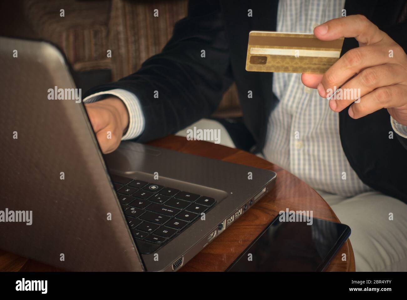 Nahaufnahme Mann in smarter Kleidung hält eine Kreditkarte kaufen Produkt online von seinem Laptop mit einem Telefon daneben auf einem Tisch zu Hause Stockfoto