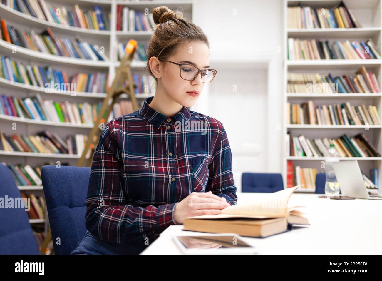 Schulmädchen in einem karierten Hemd blättert durch ein Buch, während sie in einem Bibliotheksraum sitzt. Junge Frau kam in die Bibliothek, um sich auf die Hochschulprüfung vorzubereiten. G Stockfoto