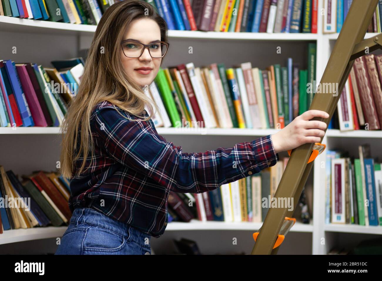 Ein hübscher Student steht auf einer Stehleiter in der Bibliothek, um das richtige Buch zu finden. Frau in der öffentlichen Bibliothek Stockfoto