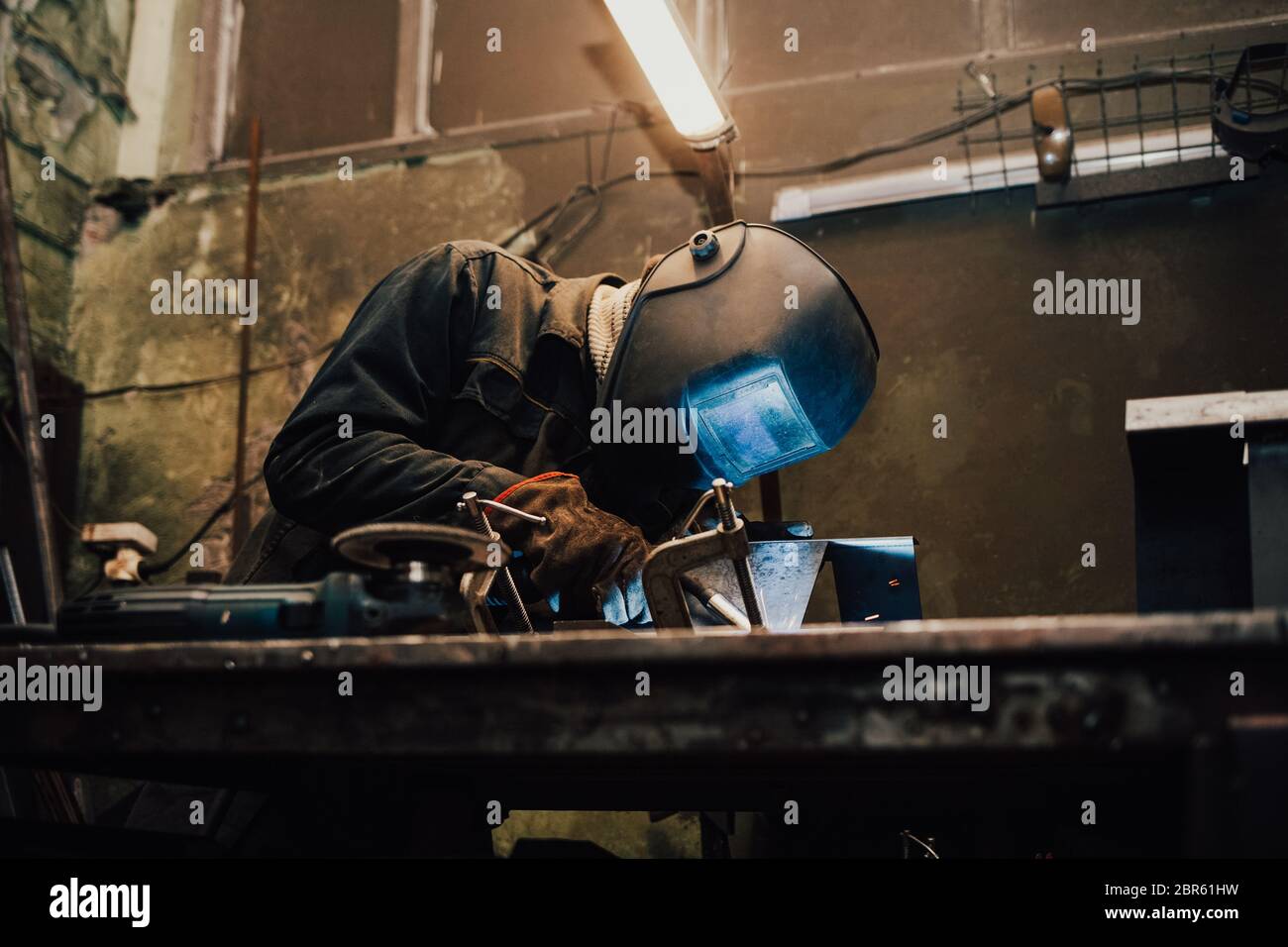 Arbeiter in einer Schweißmaske arbeitet in einer Werkstatt für das Schweißen von Eisen. Der Mensch stellt Eisenprodukte her. Guy arbeitet mit einem Schweißgerät, das eine Maske verwendet, um sein Geld zu schützen Stockfoto