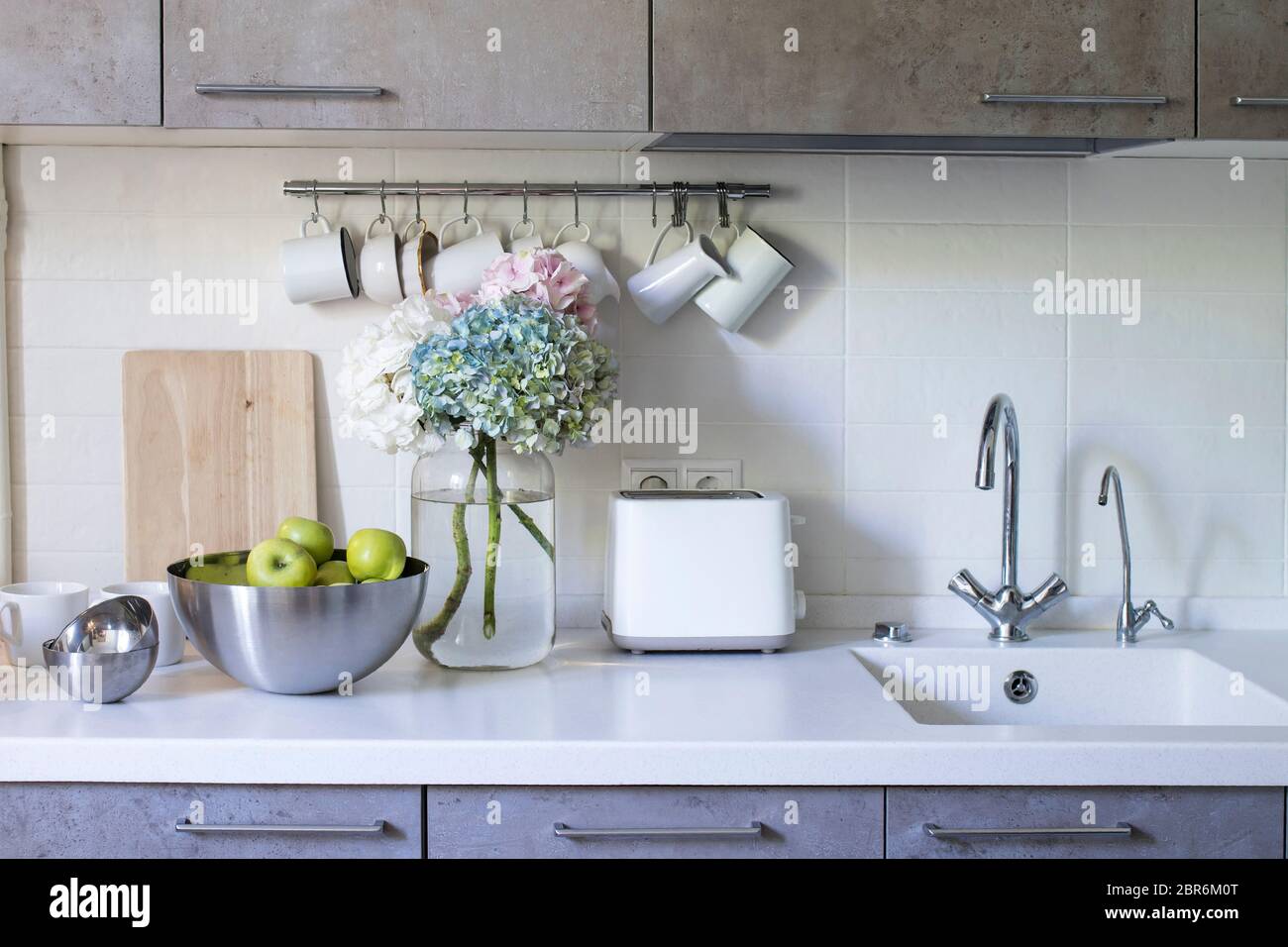 Küche in beigefarbenen Tönen. Becher und Milchkännchen hängen an Haken. Waschbecken mit Wasserhähnen. Stahlschale mit grünen Äpfeln. Bouquet von bunten Hortensien in einem Glas Stockfoto