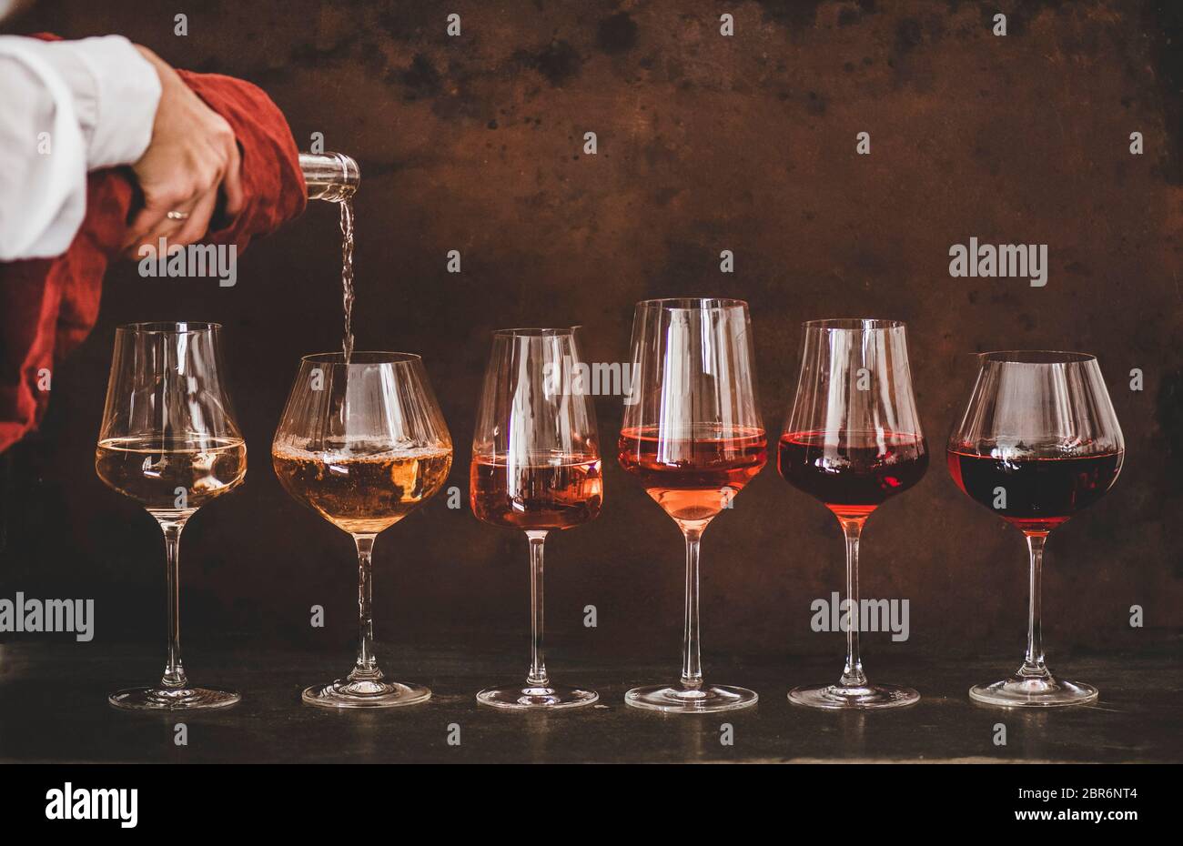 Nuancen von Roséwein in Gläsern, die in einer Linie von hellen bis dunklen Farben platziert sind und Frauen Hand Gießen Wein von Flasche zu Glas, rostig brauner Backgro Stockfoto