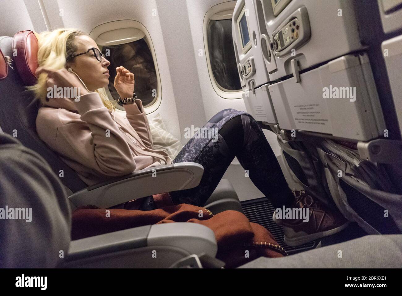 Sportlich legere junge blonde kaukasische Dame, die während der Fahrt mit dem Flugzeug am Fenster einen Film sieht. Kommerzieller Transport mit Flugzeugen Stockfoto