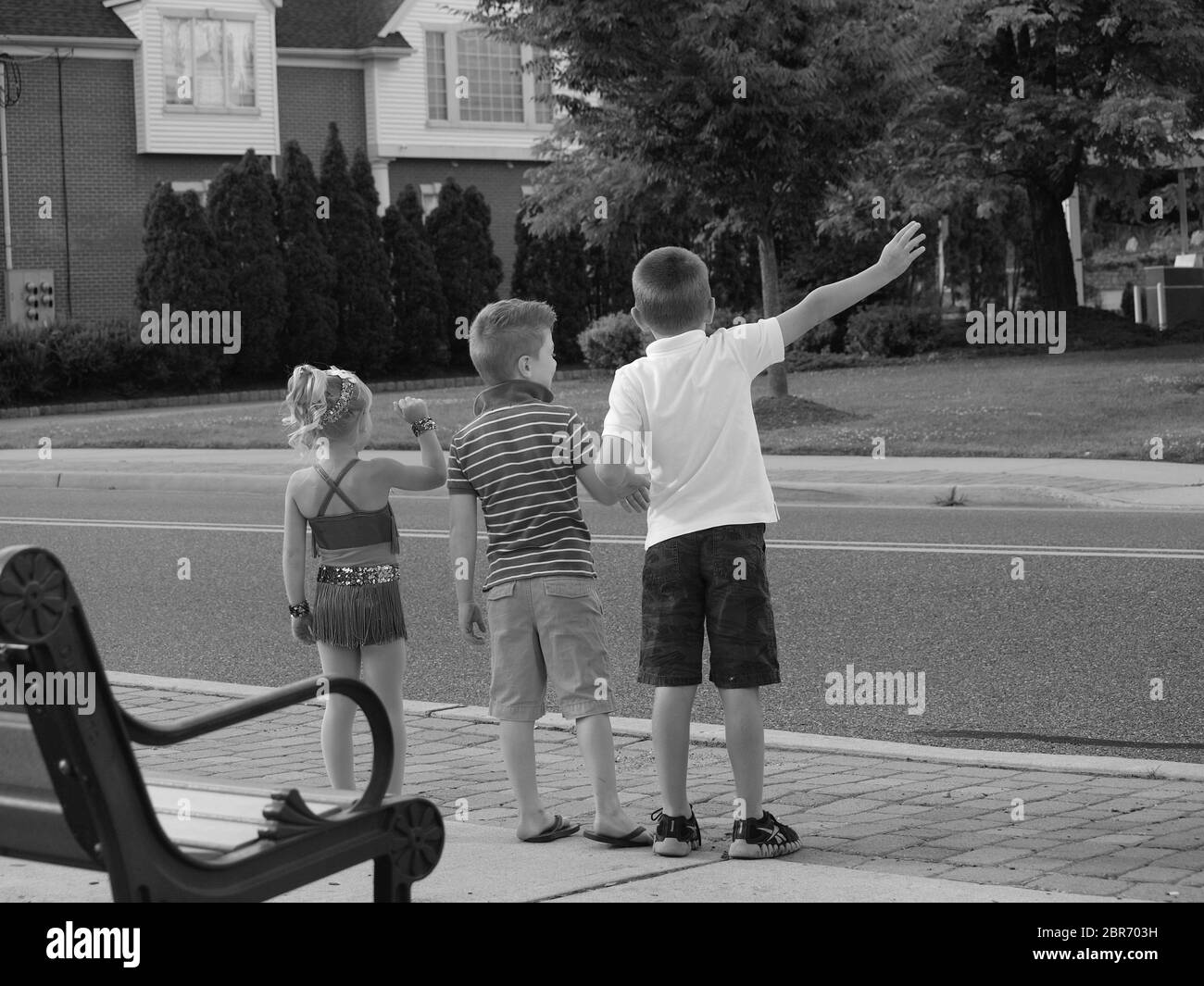 Drei Kinder versuchen Passanten entlang einer Straße in ihrer Heimatstadt anzuwinken. In schwarz und weiß zeigt eine einfachere Spaßzeit. Stockfoto