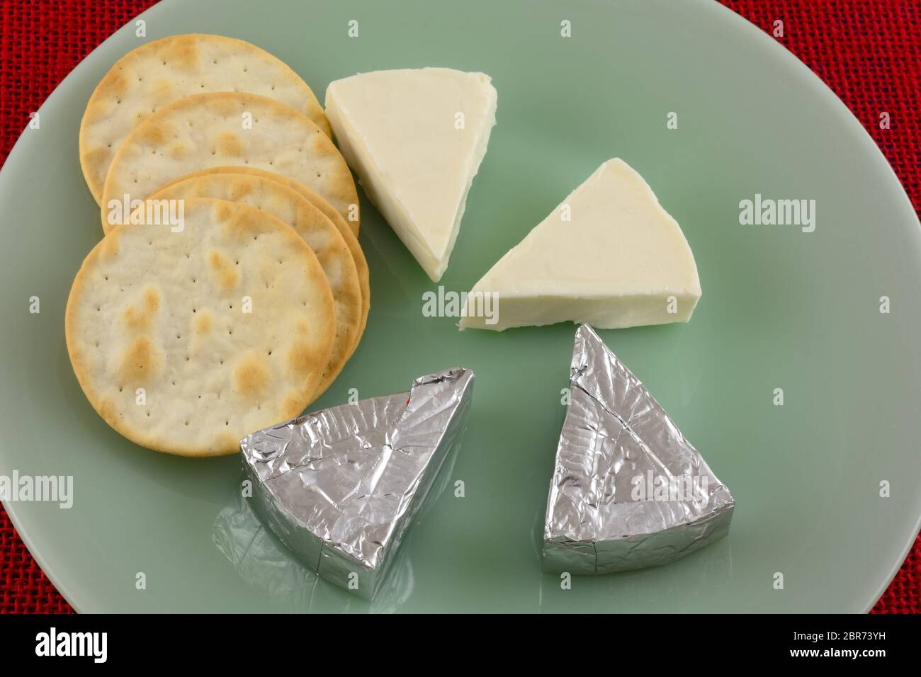 Brie Käse Mini Scheiben ausgepackt und in Aluminiumfolie verpackt und Tischwasser Cracker auf grüner Platte verpackt Stockfoto