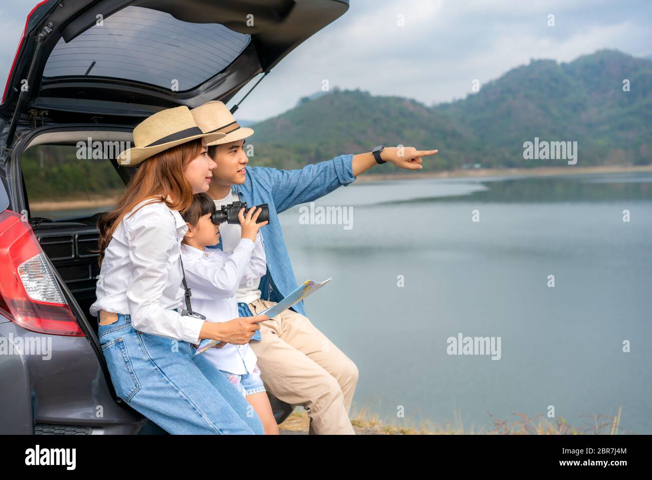 Porträt der asiatischen Familie, die im Auto sitzt, mit dem Vater, der auf Ansicht zeigt, und Mutter, die Karten mit Tochter hält, die durch das Binokul schöne Landschaft sieht Stockfoto