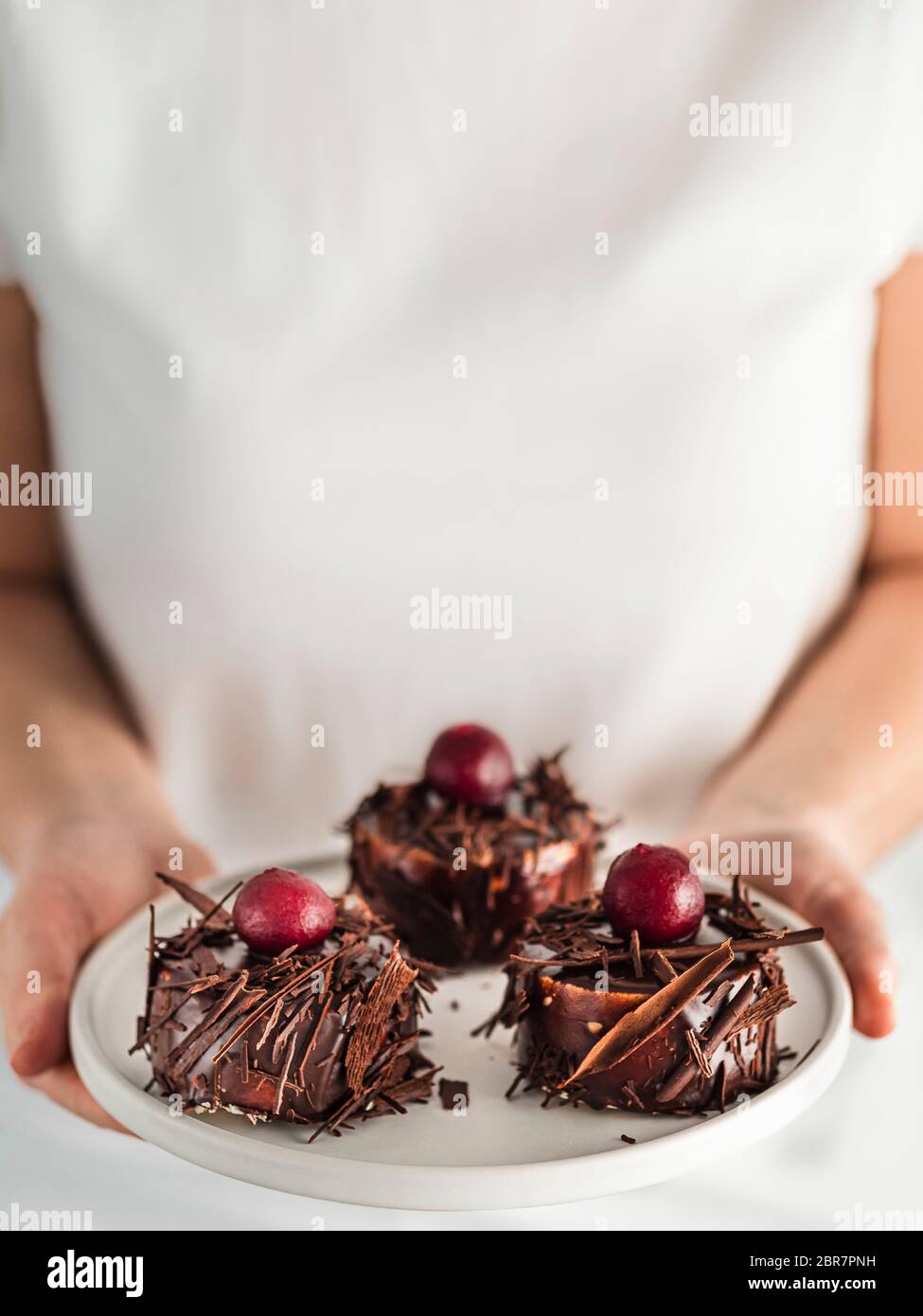 Frau in weißem T-Shirt hält Teller mit drei Mini-schwarzen Wald Kuchen mit Kirsche und Schokolade Scherben. Vertikal. Kopieren Sie den Bereich oben Stockfoto