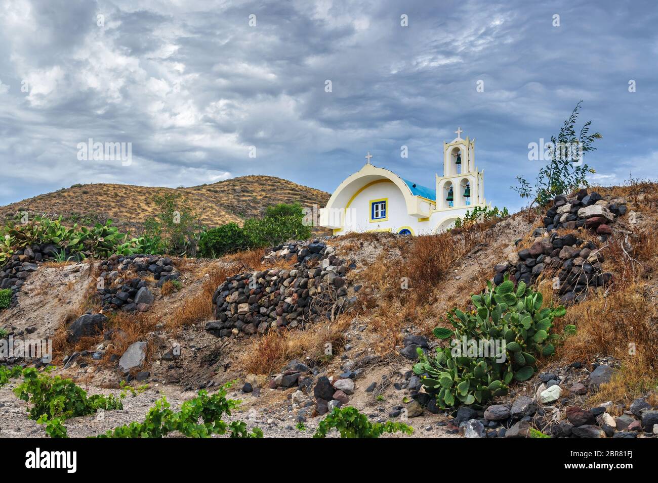 Typische Landschaft der Insel Santorini an einem Sommer bewölkten Tag. Auf dem Hügel im Dorf Akrotiri befindet sich eine traditionelle weißblaue orthodoxe Kirche Stockfoto