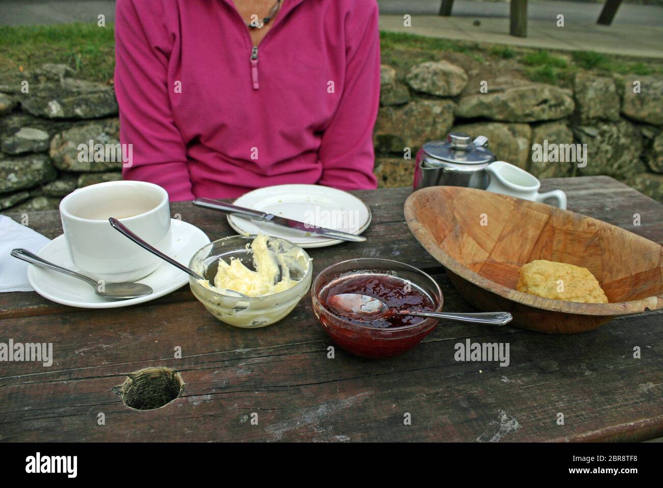 Eine nicht identifizierbare Person saß an einer Picknickbank, nachdem sie einen Cream Tea in einem Pub-Garten gegessen hatte. Tee, Scone, Sahne und Marmelade auf dem Tisch mit Besteck. Stockfoto