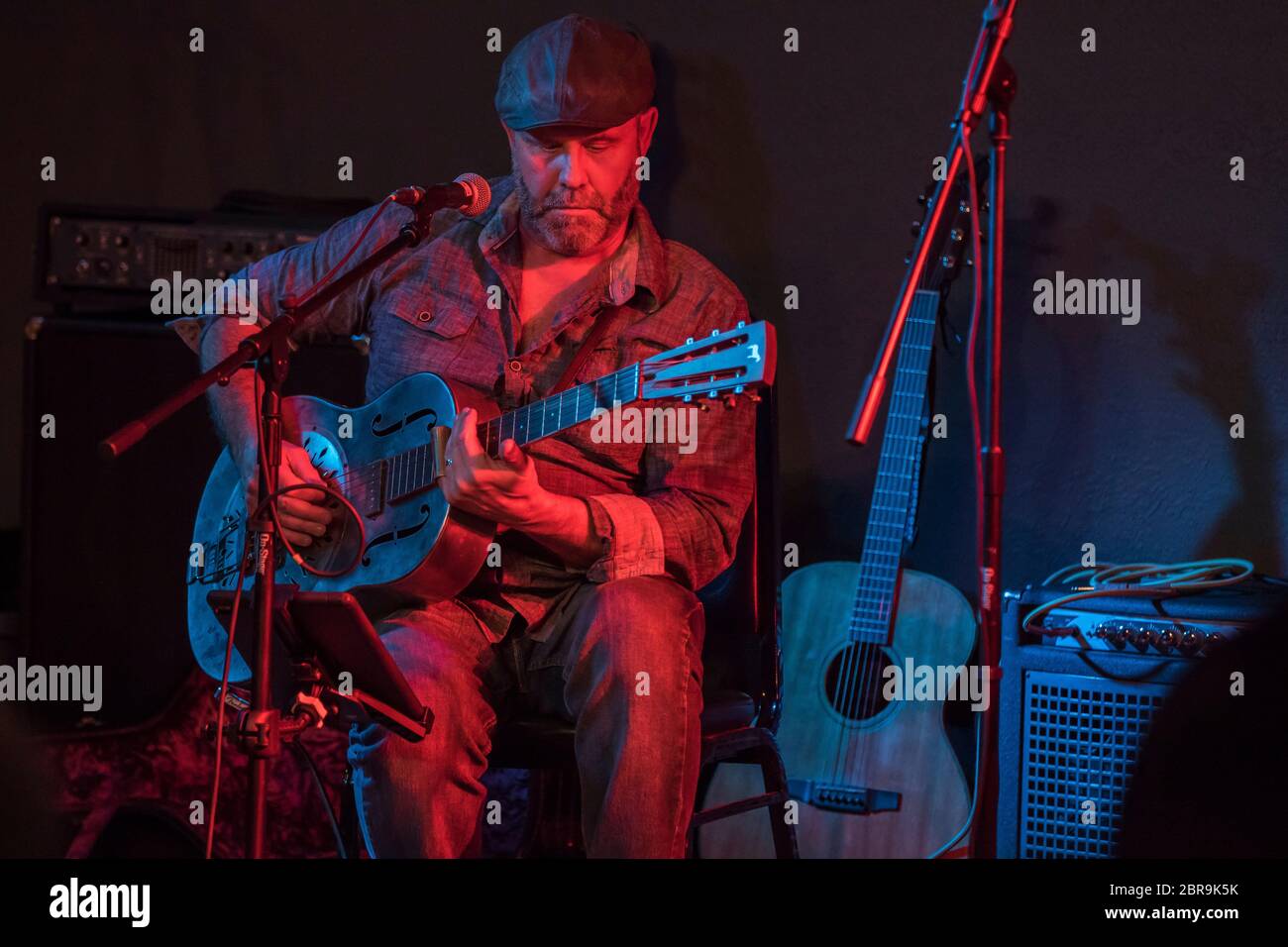 Männlicher Folksänger, der sitzt, während er singt und Resonator Gitarre spielt, in einem Indoor-Konzert. Stockfoto