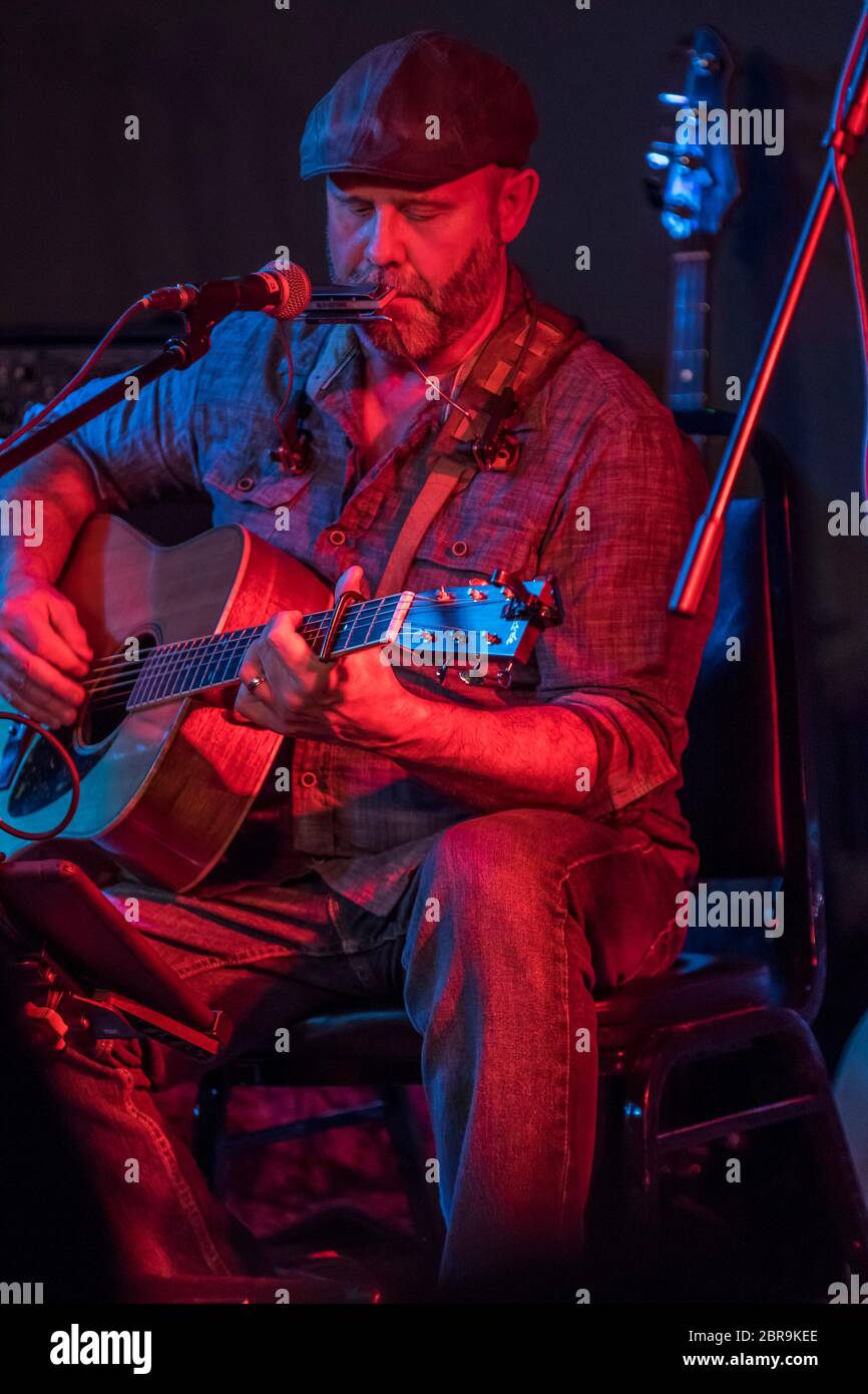 Männlicher Folksänger, der sitzt, während er singt und Gitarre spielt, in einem Indoor-Konzert. Stockfoto