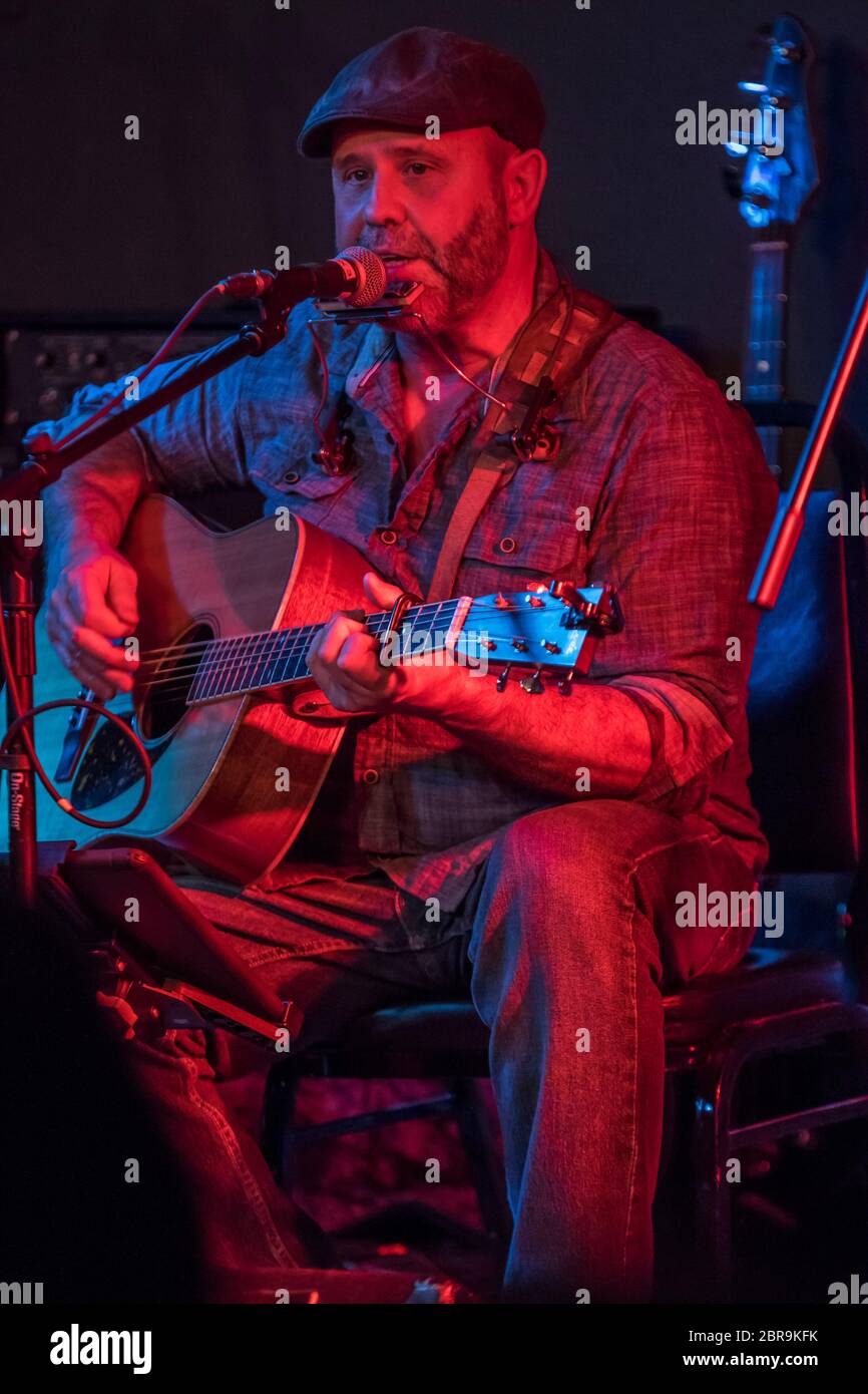 Männlicher Folksänger, der sitzt, während er singt und Gitarre spielt, in einem Indoor-Konzert. Stockfoto