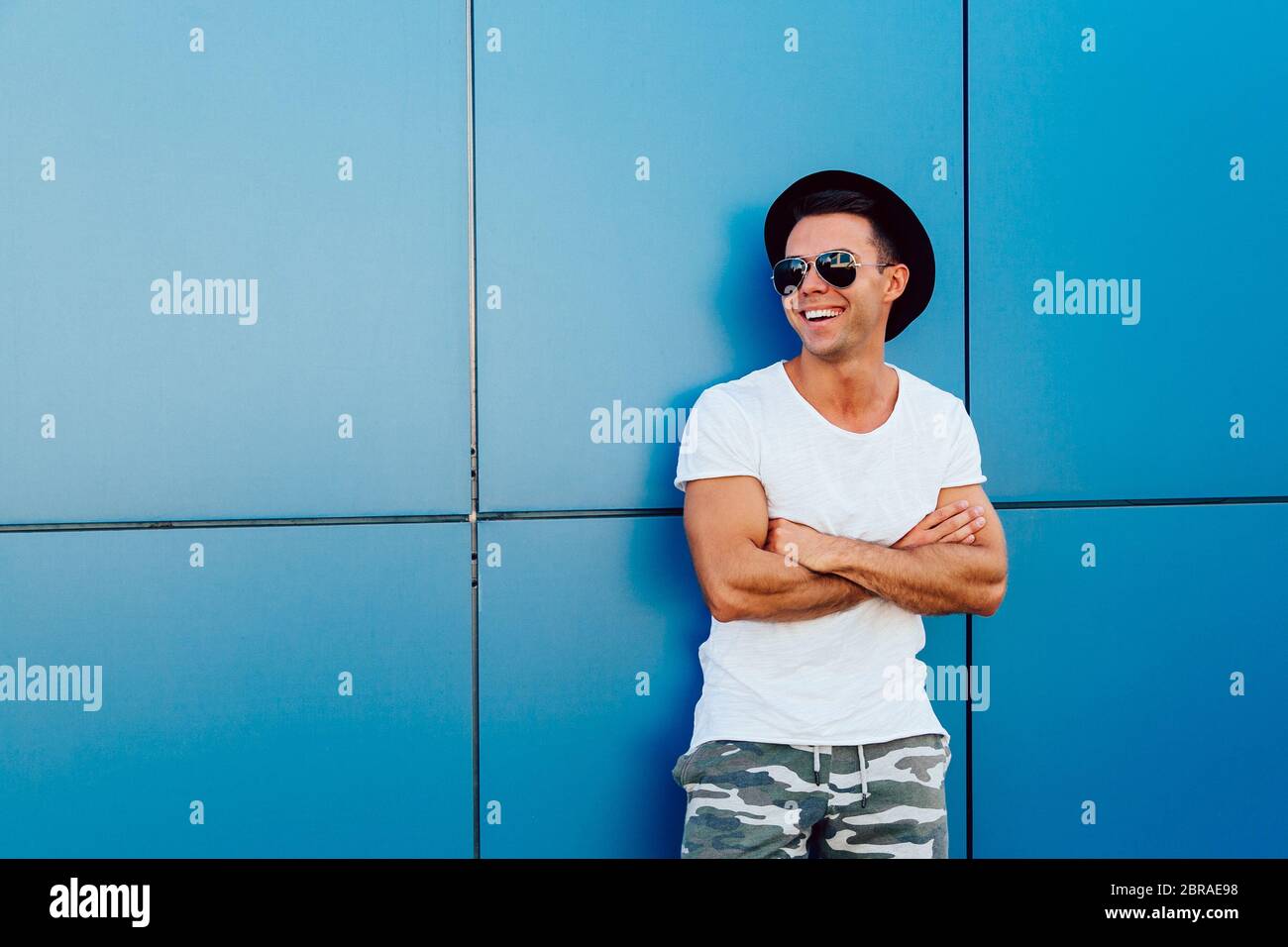 Hübscher junger Mann in Sonnenbrillen ihre Arme Kreuzung beim Stehen auf blaue Wand Hintergrund mit Kopie Raum, lächelnd und Wegsehen. Gekleidet in T-Sh Stockfoto