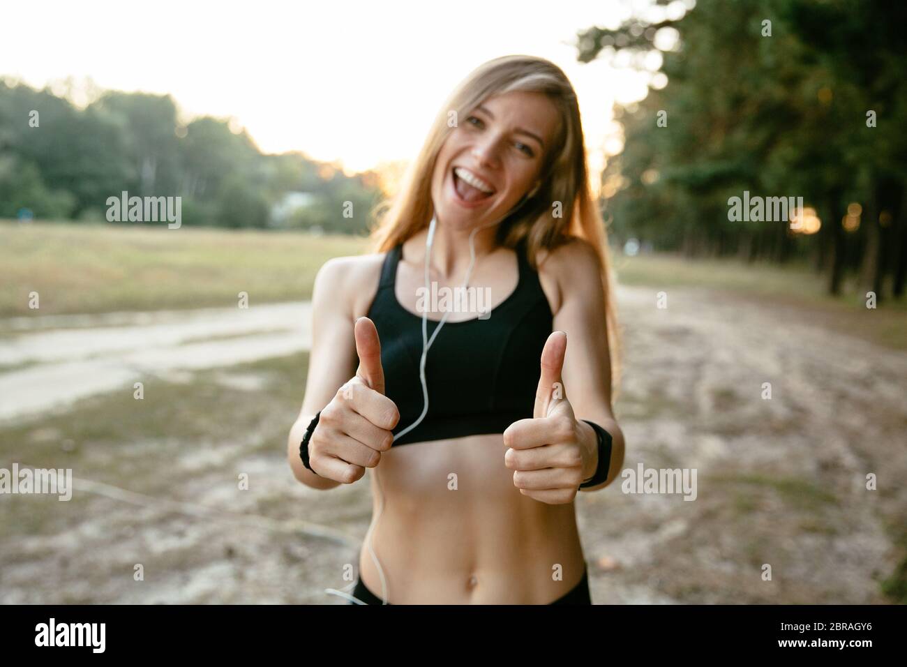 Sommer Portrait von sportliche Mädchen im Tank Top, Daumen hoch, fröhlich lächelnd und mit Blick auf die Kamera. Trainieren Sie außerhalb. Stockfoto