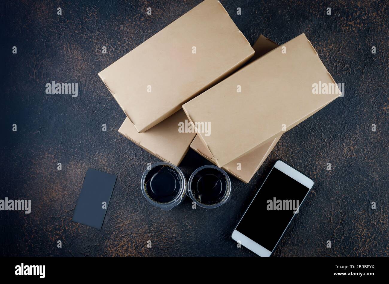 Einweg-Kraftpapier-Lebensmittelboxen für Sushi-Brötchen, Glas mit Sauce und Essstäbchen auf dunkel Stockfoto