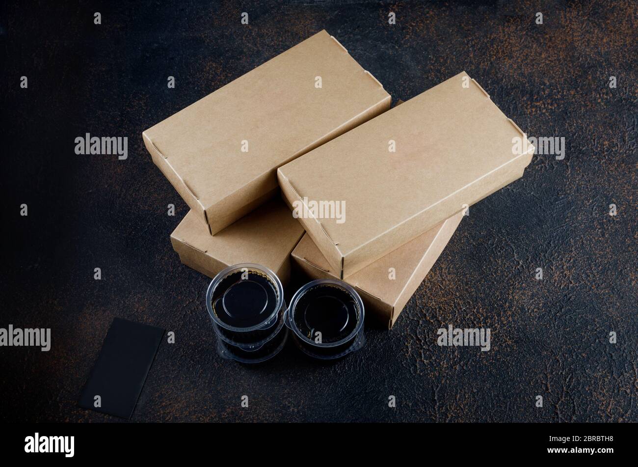 Einweg-Kraftpapier-Lebensmittelboxen für Sushi-Brötchen, Glas mit Sauce und Essstäbchen auf dunkel Stockfoto