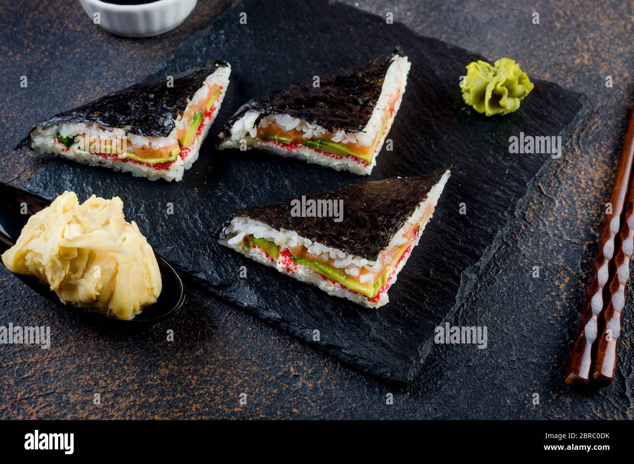 Leckere Sushi-Sandwiches mit Lachs auf schwarzem Steinteller mit Sojasoße, Ingwer, Wasabi Stockfoto