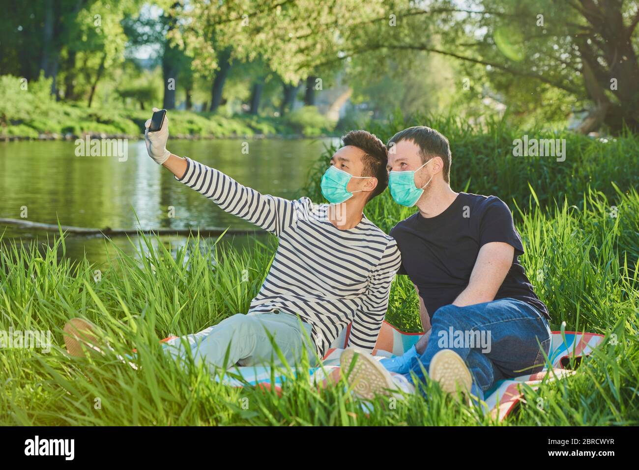 Zwei homosexuelle Männer mit Gesichtsmasken sitzen auf einer Wiese am Fluss, machen ein Selfie, Corona-Krise, Donau, Bayern, Deutschland Stockfoto