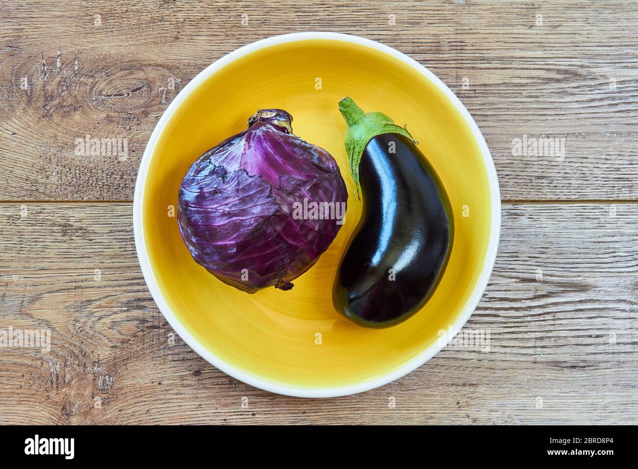 Kleiner violetter Kopf aus Rotkohl und fast schwarzer Aubergine auf einer gelben Platte auf einer Holzplatte Stockfoto