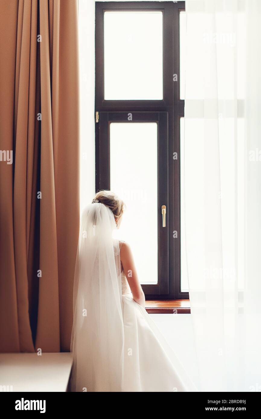 Braut in Hochzeitskleid stehend am Fenster im Zimmer mit Blick aus Stockfoto