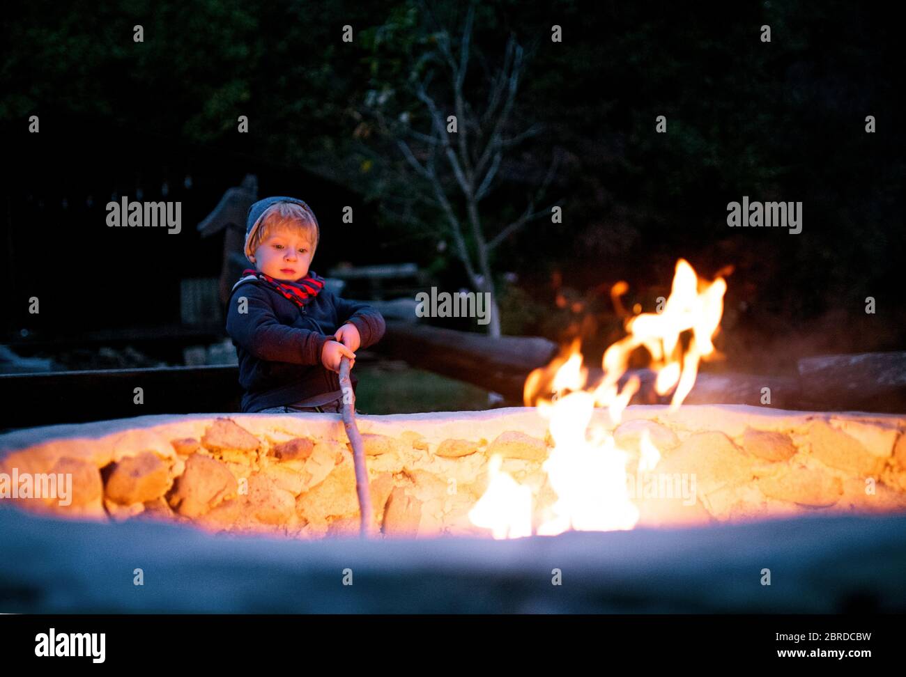 Ein Kleinkind Junge steht und spielt am offenen Feuer im Garten im Sommer. Stockfoto