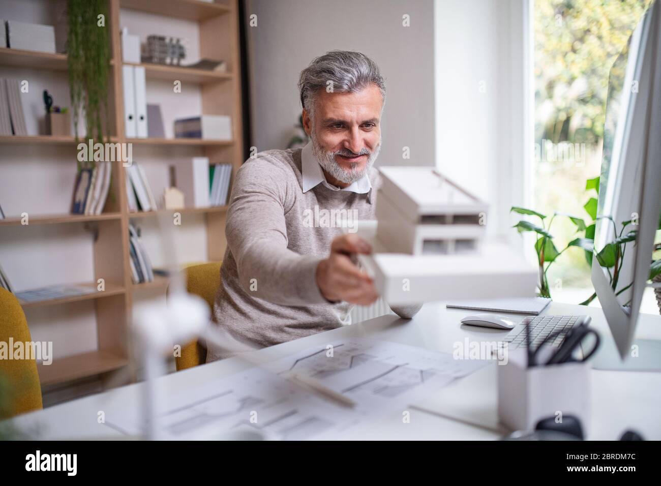 Architekt mit Modell des Hauses sitzt am Schreibtisch im Büro drinnen, arbeiten. Stockfoto