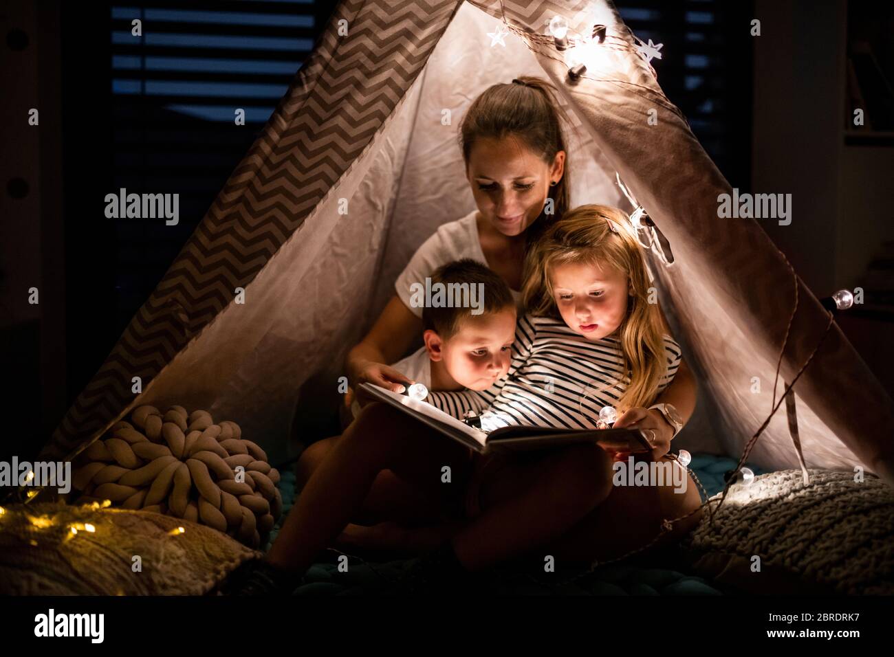 Mutter mit Kindern, die drinnen im Schlafzimmer sitzen und ein Buch lesen. Stockfoto