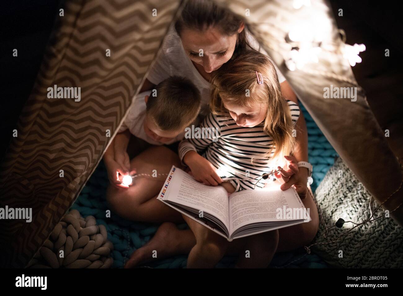 Mutter mit Kindern, die drinnen im Schlafzimmer sitzen und ein Buch lesen. Stockfoto