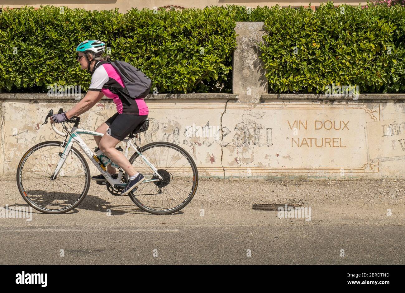 Radfahrerin, die an der alten Dorfmauer mit Weinbauschild in Rasteau in Südfrankreich vorbeifährt Stockfoto