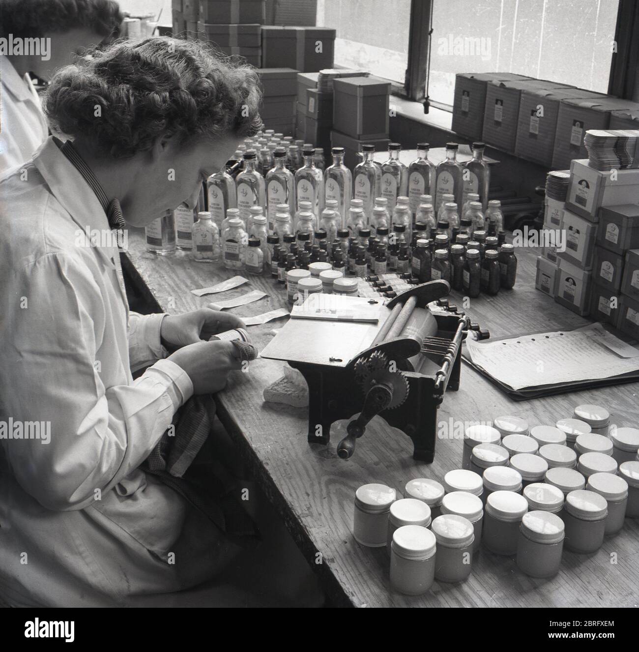 1950er Jahre, historische, weibliche Arbeiterin in einem weißen Mantel, die an einer Holzwerkbank sitzt und kleine Flaschen mit Medikamenten mit einer kleinen Handmaschine beschriftet, England, Großbritannien. Stockfoto