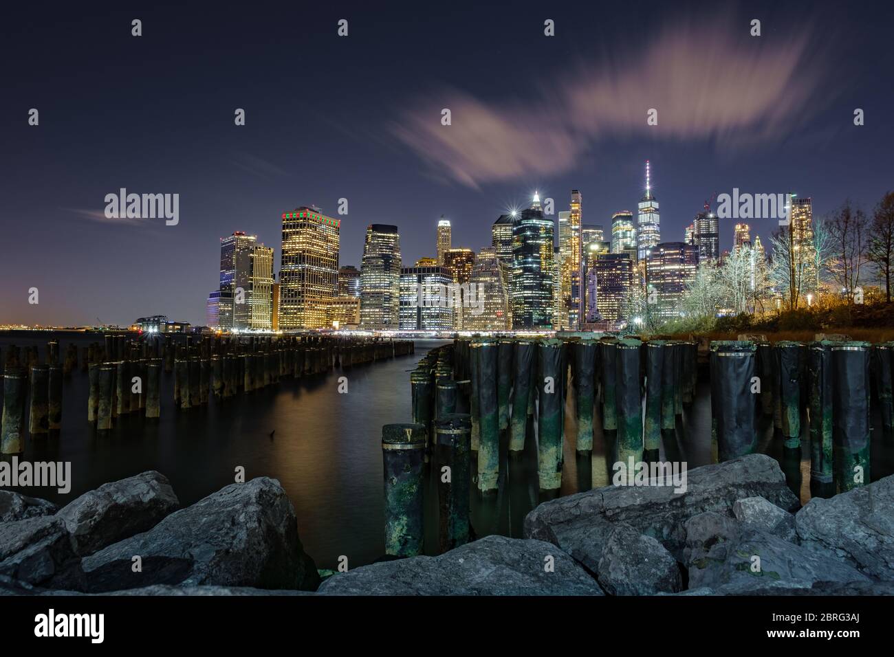 Eine lange Exposition des Finanzdistrikts von Manhattan, aufgenommen vom Brooklyn Bridge Park mit den Polen im Wasser, New York, USA Stockfoto
