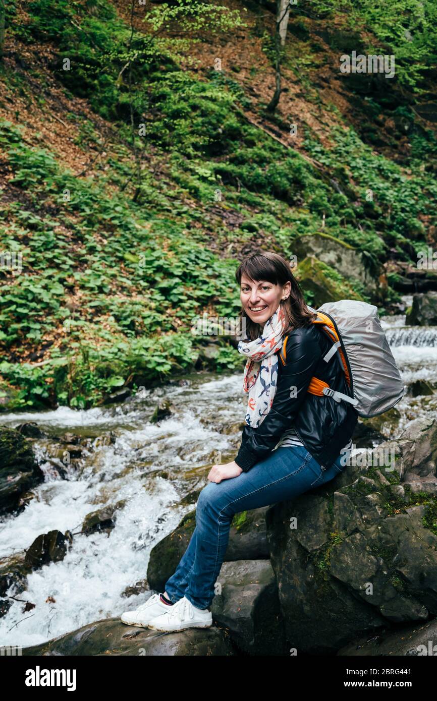 Fröhliches lächelndes Touristenmädchen in blauen Jeans, gestreiftes Hemd und schwarze Jacke mit Rucksack in der Nähe des kalten frischen Flusses am Wasserfall Shypit, Kaskade Stockfoto