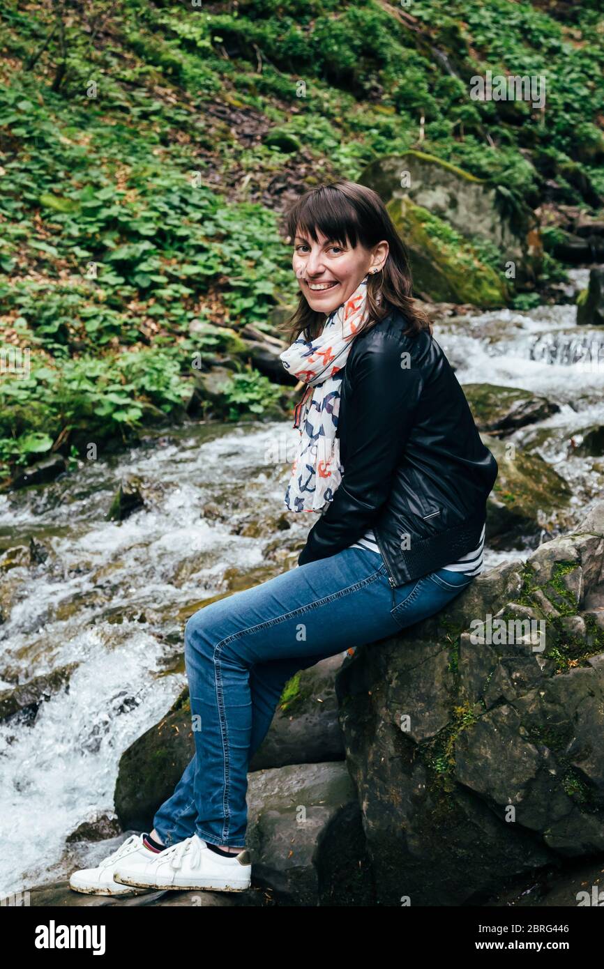 Fröhliches lächelndes Touristenmädchen in blauer Jeans, gestreiftem Hemd und schwarzer Jacke nahe dem kalten frischen Fluss am Wasserfall Shypit, Kaskade in Pylypets innen Stockfoto