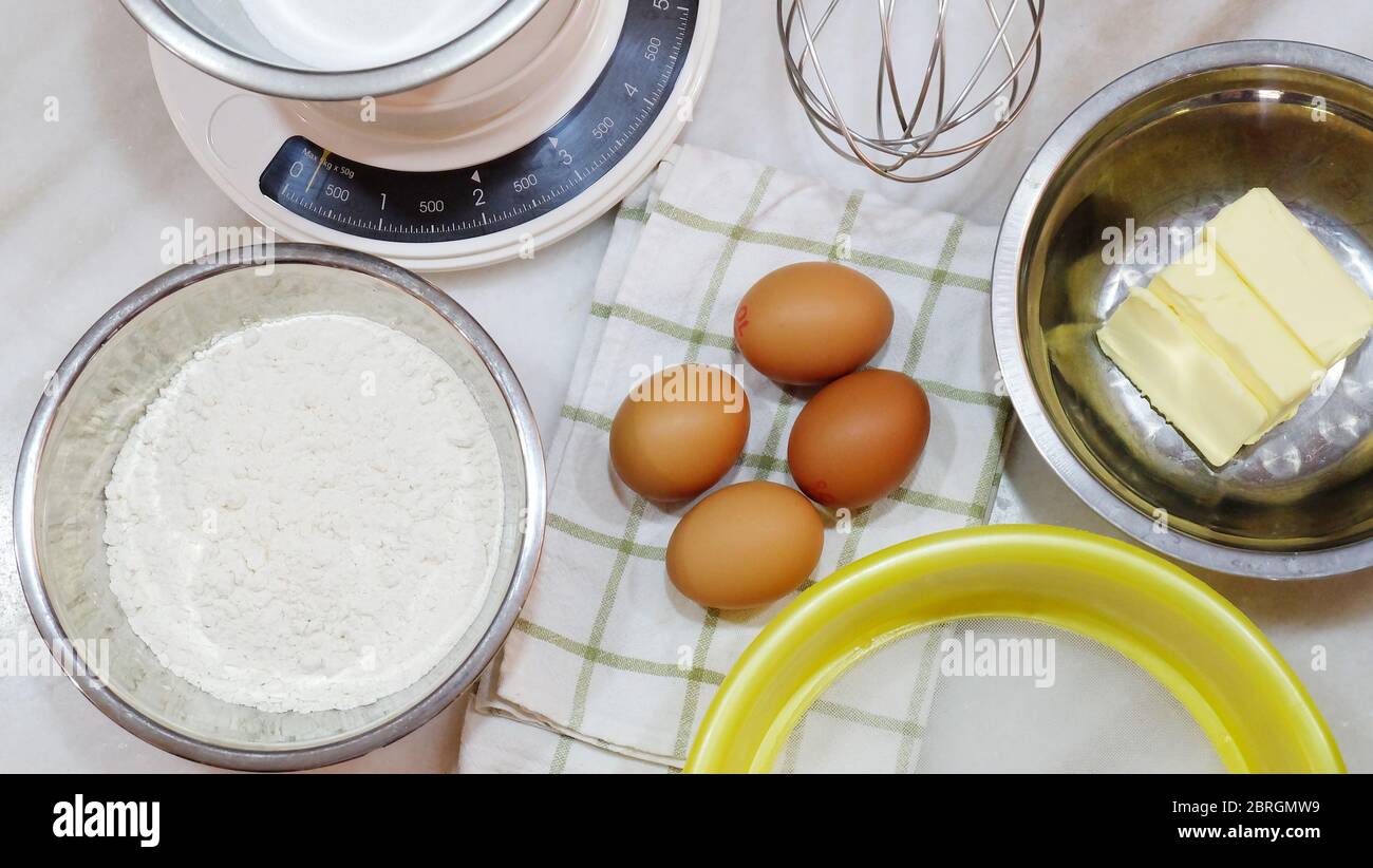 Backzutaten aus braunen Eiern, Weißmehl, Butter, Mehlsichter, Küchenwaage und einem Metallbesen, auf einer Tischplatte platziert. Stockfoto