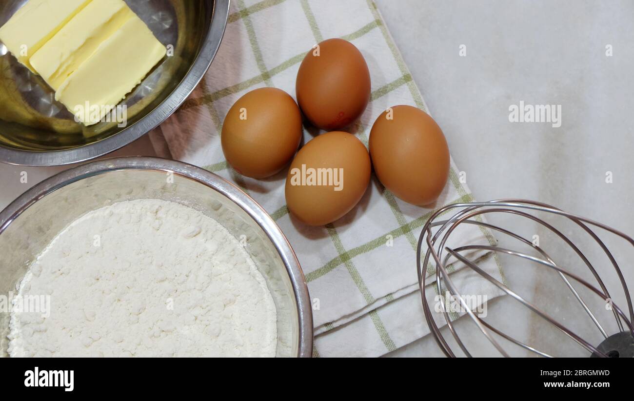 Backzutaten aus braunen Eiern, weißem Mehl, Butter und einem Metallbesen, auf einer Tischplatte platziert. Stockfoto