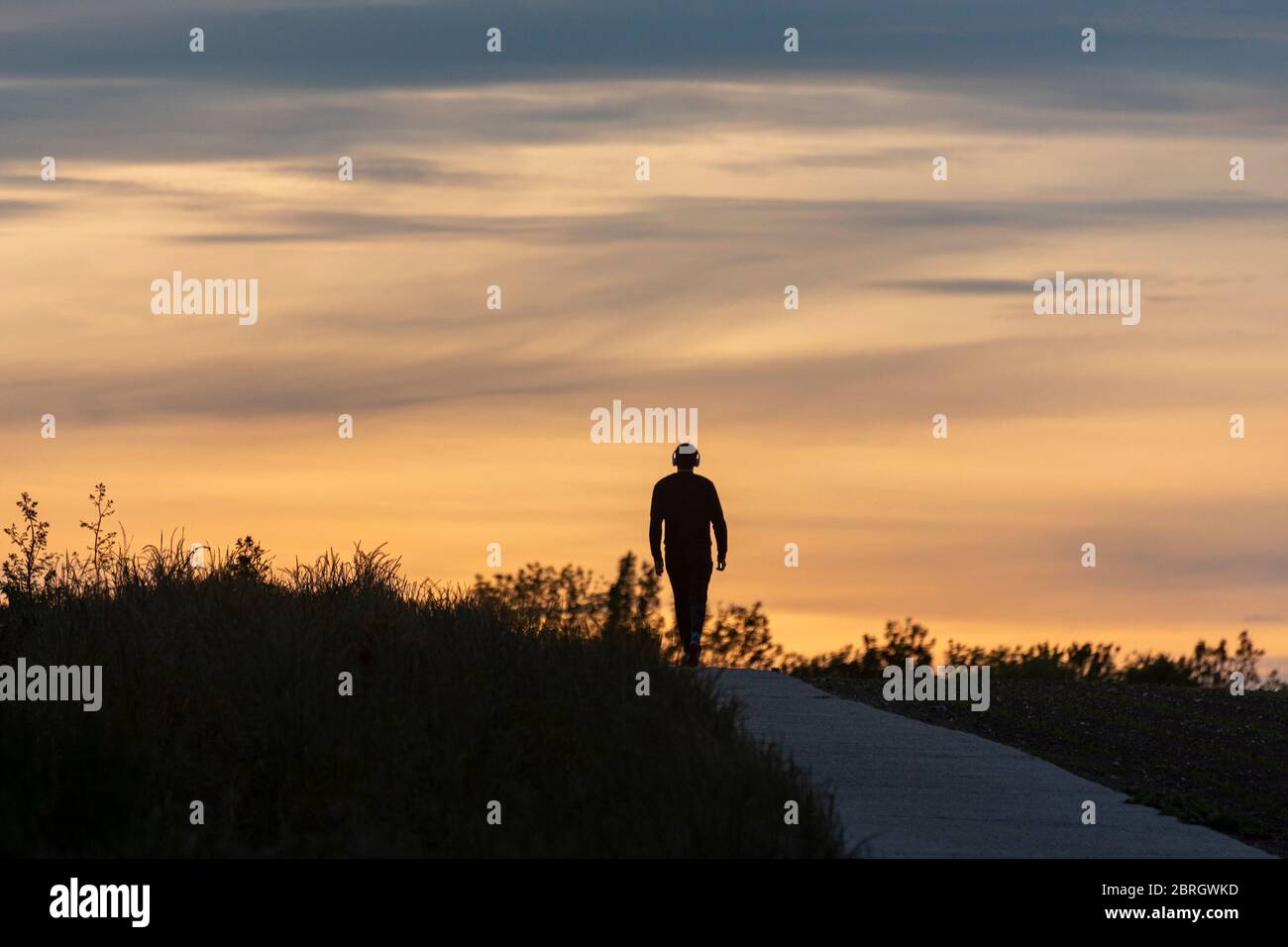 Die Silhouette eines Mannes erreicht den Horizont auf einem Pfad, während seines Abendspaziergangs, mit Kopfhörern an. Stockfoto