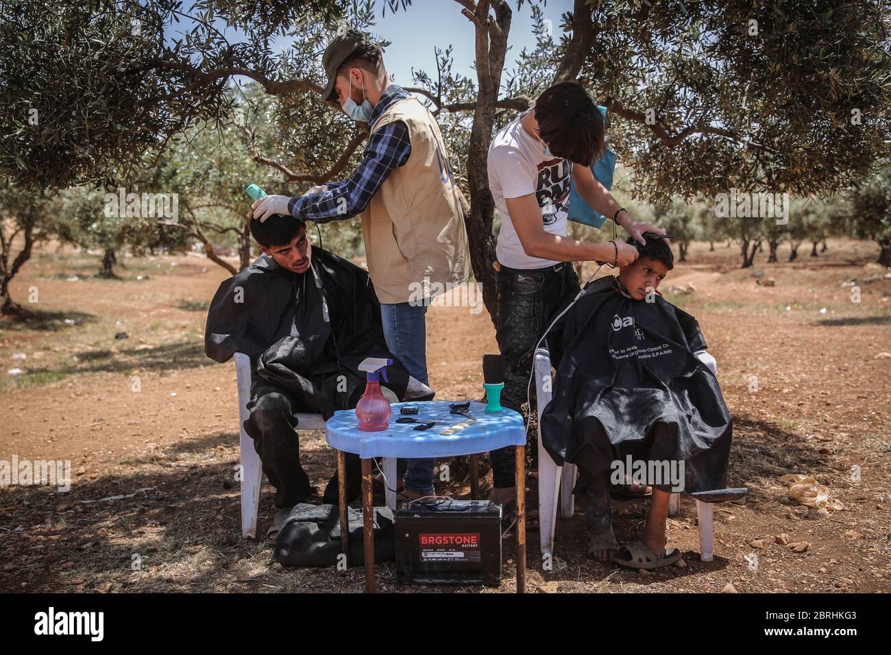 Kah, Syrien. Mai 2020. Ehrenamtlicher Friseur mit Gesichtsmasken gibt intern vertriebenen Kindern einen Haarschnitt vor Eid Al-Fitr in einem Flüchtlingslager im Dorf Kah in der nördlichen Idlib-Provinz nahe der Grenze zur Türkei. Eine Gruppe von 10 Friseuren eines ehrenamtlichen Teams namens "Al Helm, der Traum" bereist die Flüchtlingslager, um Kindern einen Haarschnitt zu geben und zielt in ihrer letzten Kampagne vor Eid Feiern auf mindestens 1000 Kinder. Quelle: Anas Alkharboutli/dpa/Alamy Live News Stockfoto