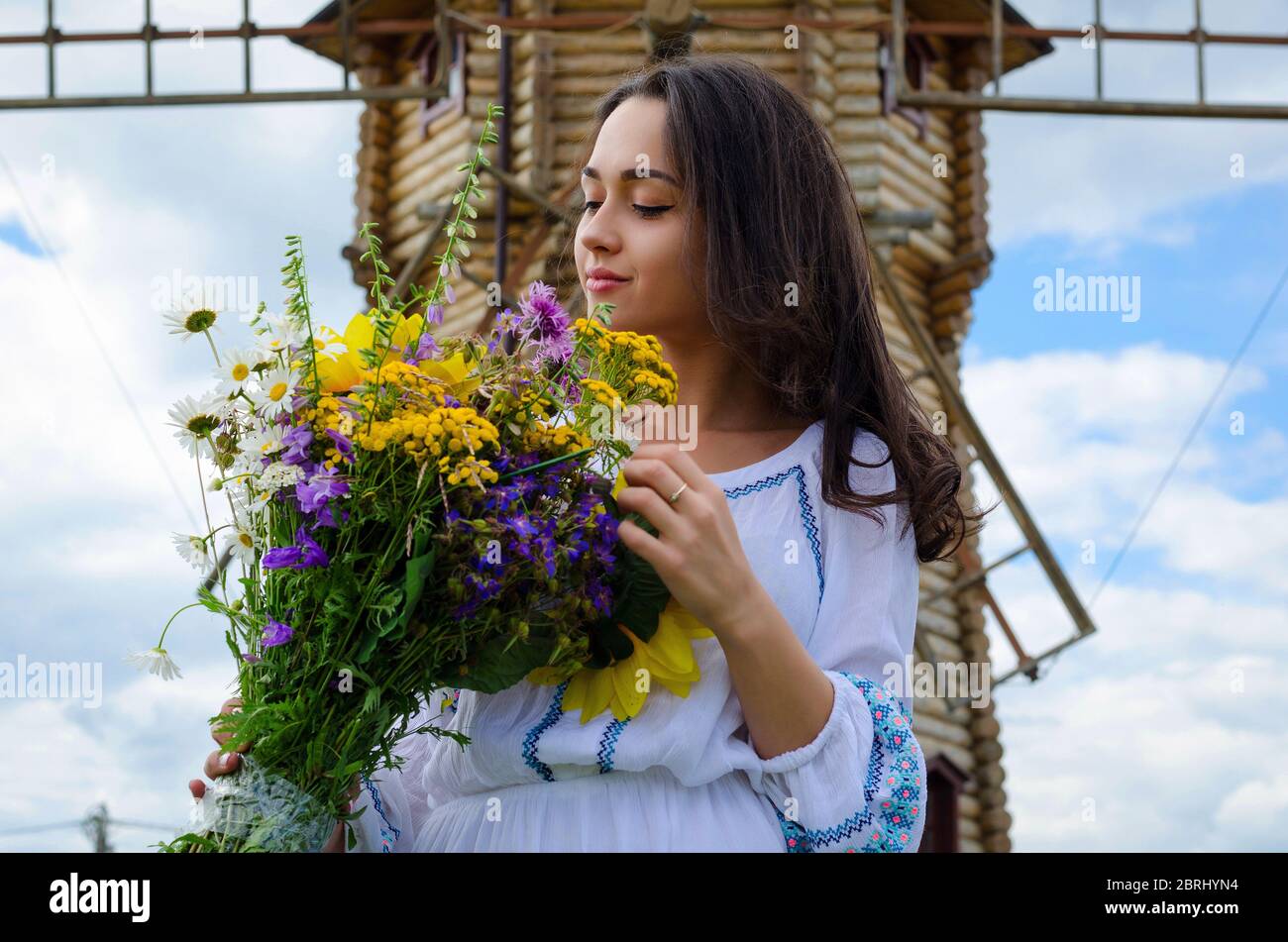 Ein junges Mädchen in einem weißen Kleid in der Nähe einer Windmühle.Mädchen in Tracht und Wildblumen Stockfoto