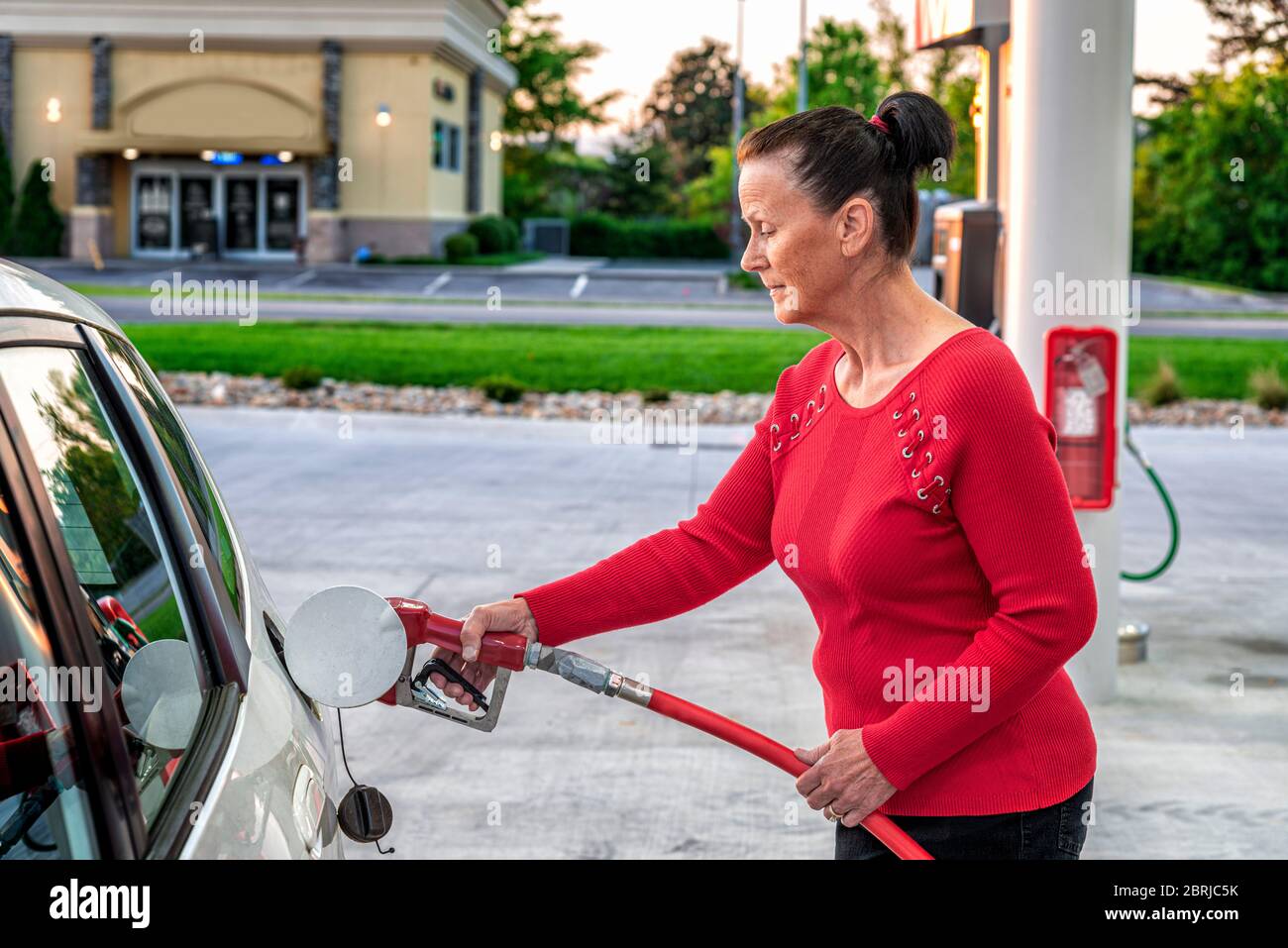 Horizontale Aufnahme einer Frau mittleren Alters mit ihren Haaren in einem Brötchen, das am frühen Morgen Gas in ihr weißes Auto pumpt. Stockfoto