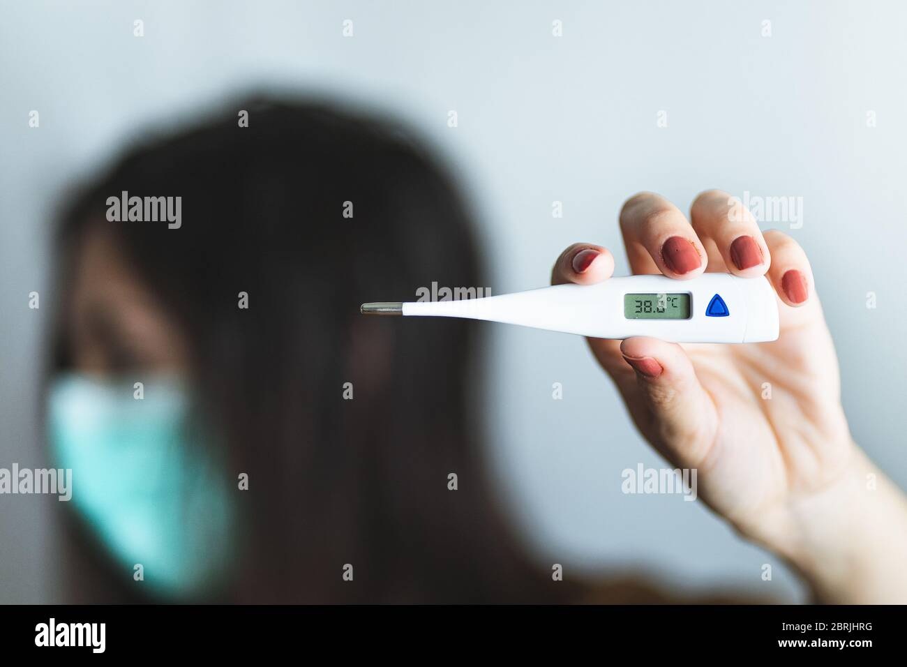 Vorderansicht der Hand einer Frau, die ein digitales Thermometer hält, das über 38 Grad Fieber anzeigt. Hintergrund der kranken Frau, die eine chirurgische Maske trägt, besorgt Stockfoto