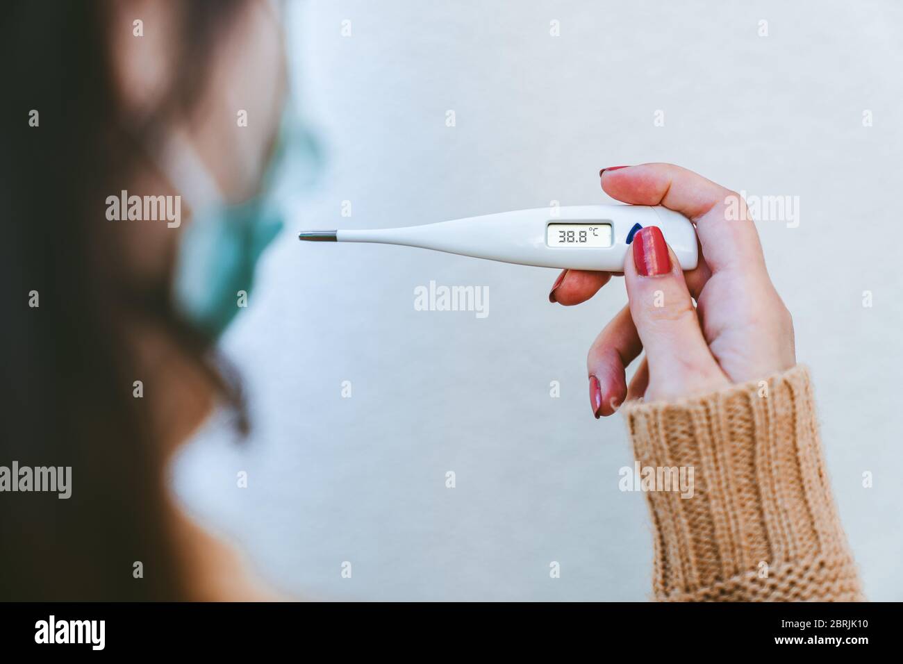 Nahaufnahme einer kranken jungen Frau, die eine chirurgische Maske trägt und ein digitales Thermometer hält, das anzeigt, dass sie über 38 Grad Fieber hat. Stockfoto
