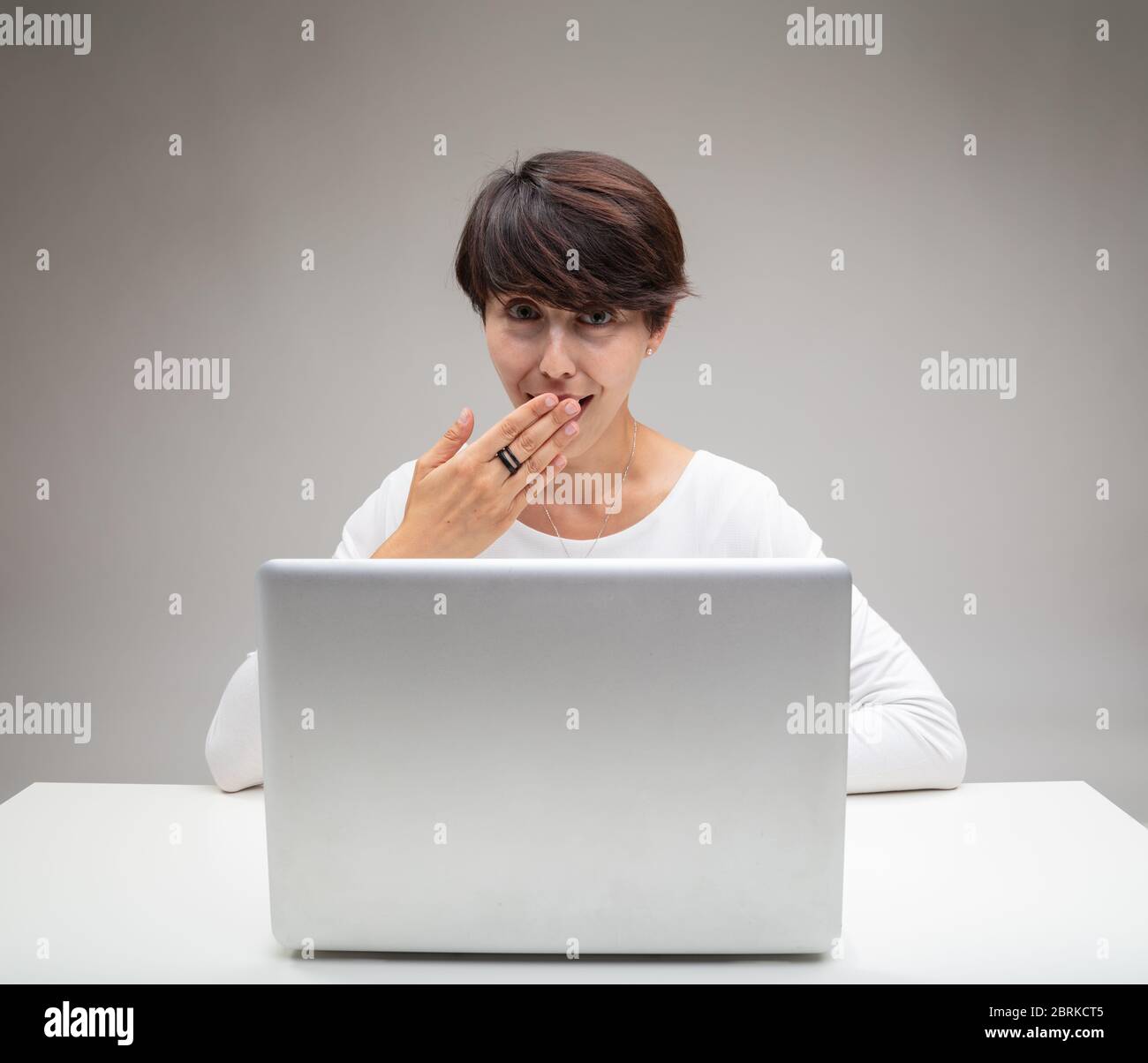 Taktvolle Frau, die ihren Mund bedeckt, während sie sich amüsiert hinter einem Laptop vor einem grauen Hintergrund sitzt Stockfoto