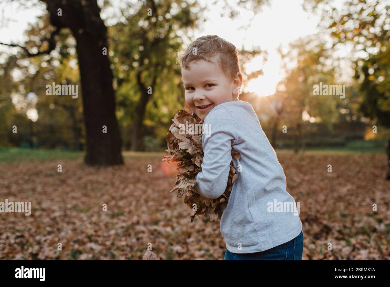 Junger Junge, der Herbstblätter hält. Nettes Kind mit einem Arm von trockenen braunen Blättern aus dem Boden lächelnd und Blick auf die Kamera. Stockfoto