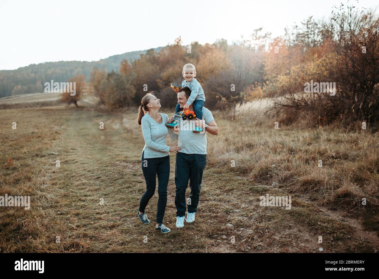 Familie zu Fuß mit Sohn im Feld. Eltern mit einem kleinen Jungen auf den Schultern des Vaters, der auf dem Land spazierend und lächelnd ist. Stockfoto