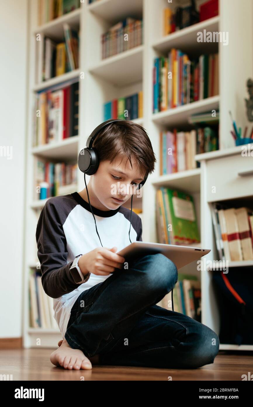 Kniender Junge lernt online zu Hause. Ein seriöser junger Student, der seine Heimarbeit mit einem digitalen Tablet macht. Stockfoto