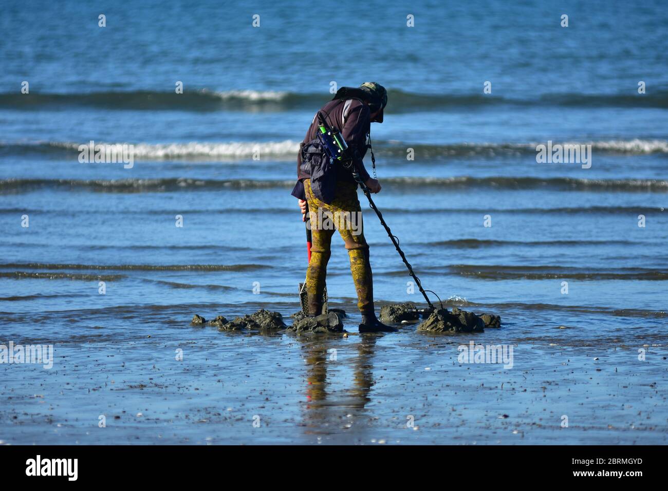 Schatzsucher in Camo Neoprenanzug Hose mit Metalldetektor und Schaufel am sandigen Ufer. Stockfoto