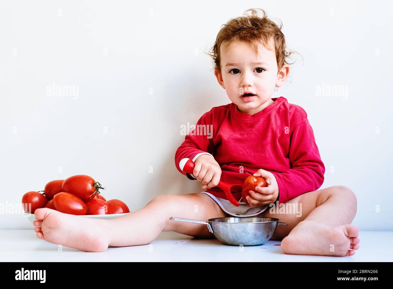 Baby sieht beim Kauen von Gemüse mit natürlichem Tomatensaft verschmutzt aus, isoliert auf Weiß. Stockfoto