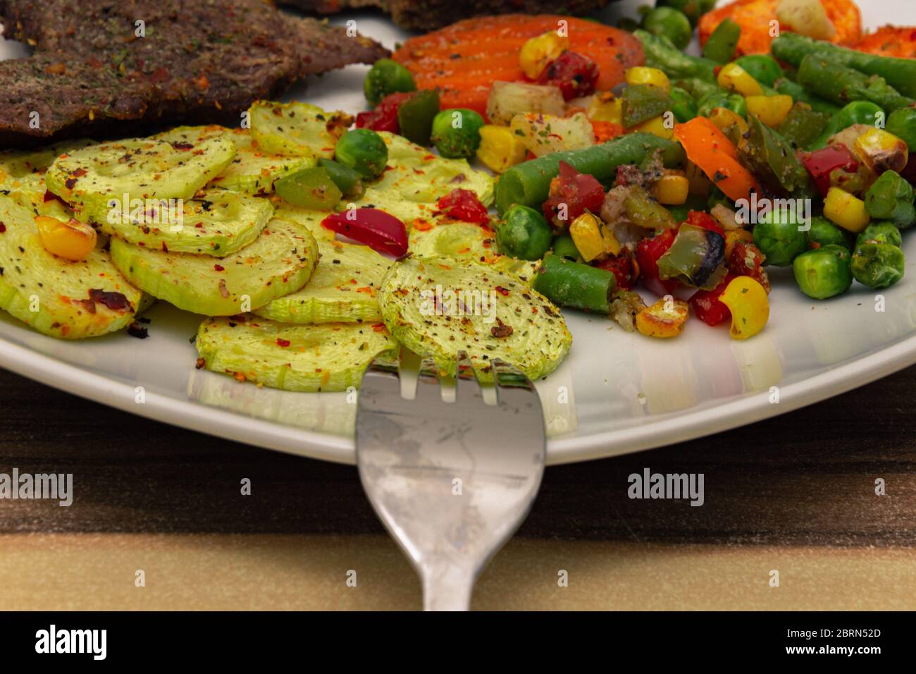 Gewürzte gekochte Zucchini neben anderem Gemüse und Fleisch auf einem weißen Teller Stockfoto