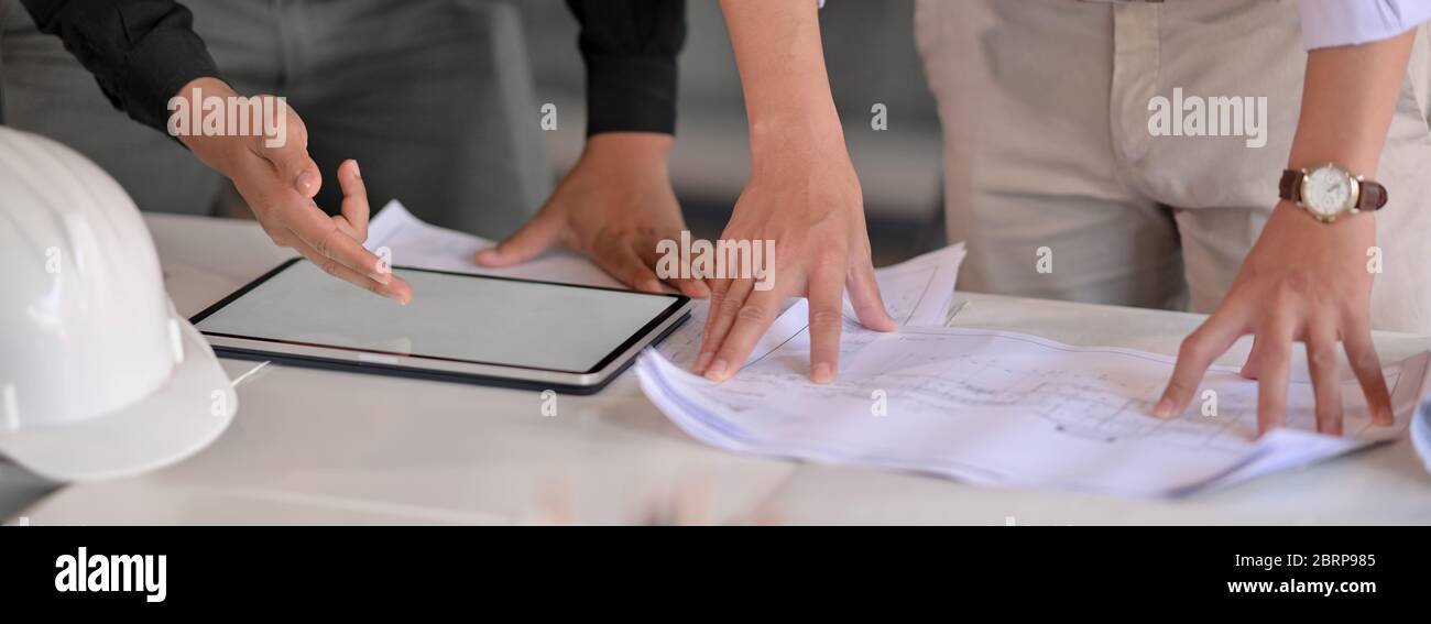 Nahaufnahme von zwei Ingenieuren, die bei ihrem Projekt mit einem Mock-up-Tablet und Papierkram auf dem weißen Tisch im Tagungsraum beraten Stockfoto