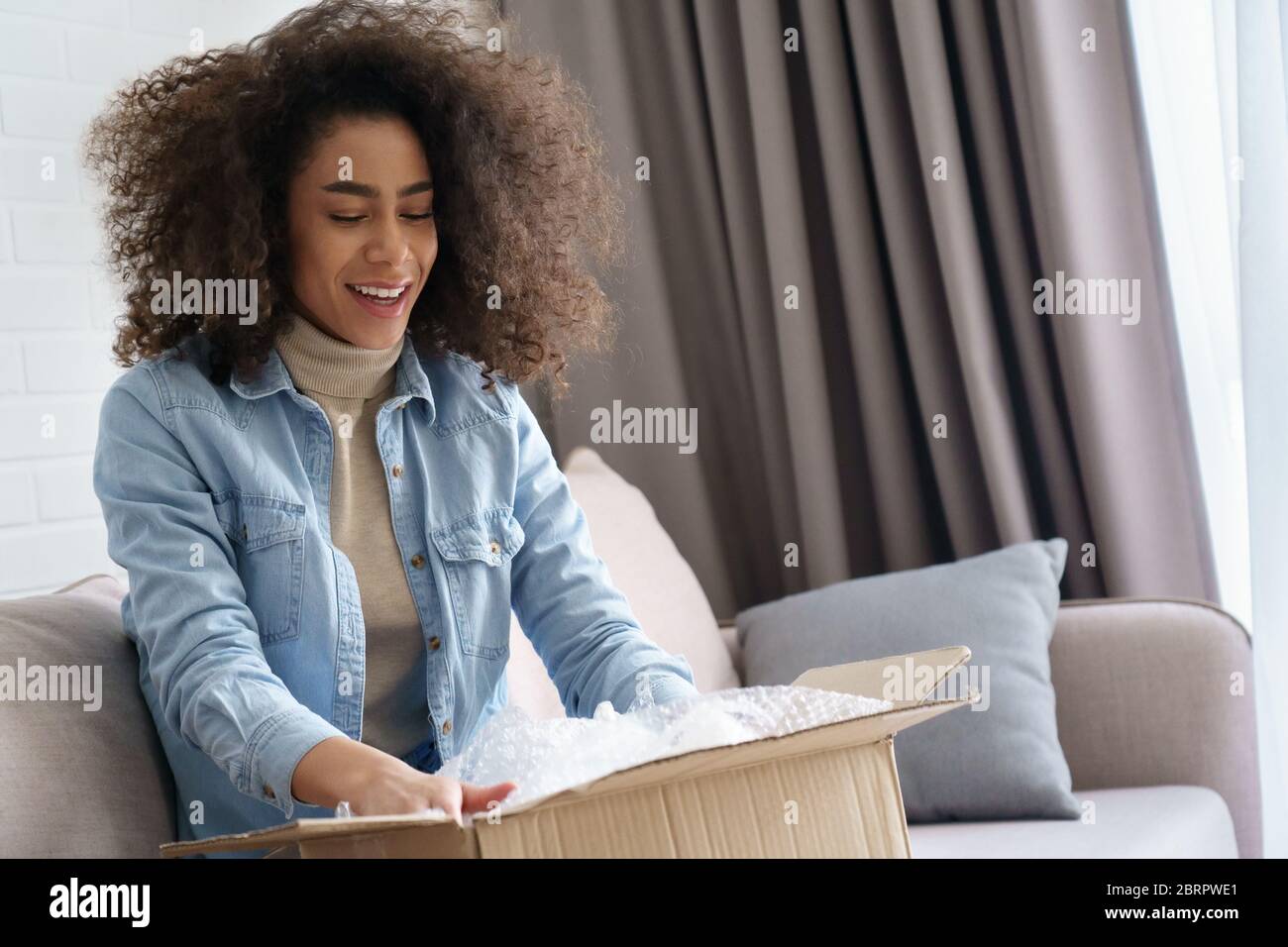 Aufgeregt afro amerikanische junge Frau Shopper Öffnung Paketkasten zu Hause. Stockfoto